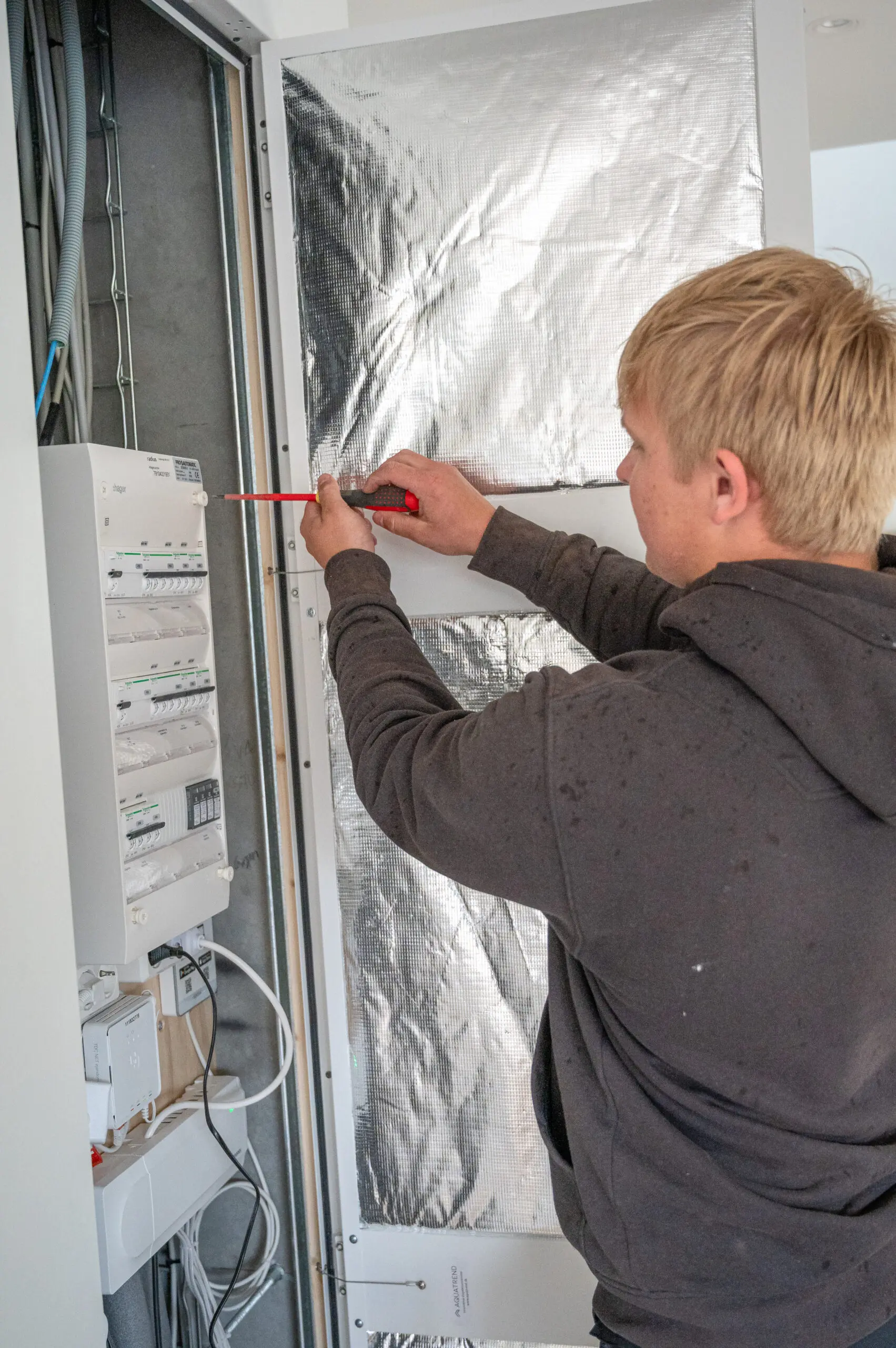 En ung elektriker fra Vejby A/S arbejder med en skruetrækker på en moderne hvid eltavle i et nybyggeri.